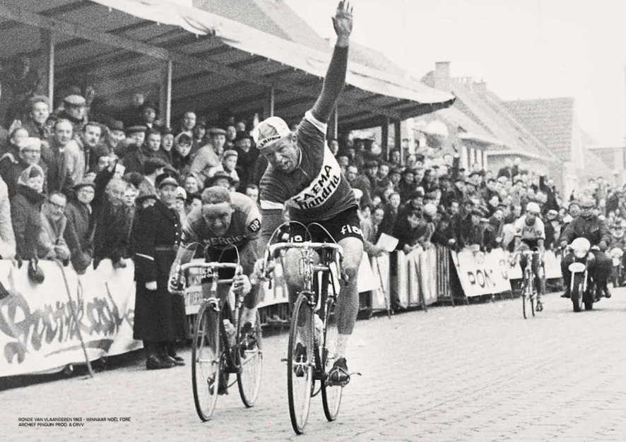 Historische zwart-witfoto van een Flandria-wielrenner die zijn arm opsteekt bij de aankomst van een koers, met toeschouwers langs het parcours